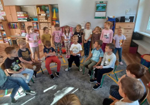Chłopcy siedzą na krzesłach w półkolu, dziewczynki uroczyście śpiewają "Sto lat"