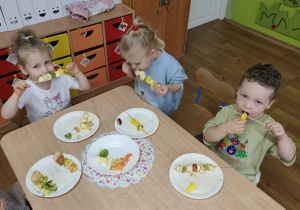 Dzieci siedzą przy stolikach i jedzą szaszłyki