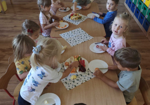 Dzieci siedzą przy stolikach i kroją owoce na sałatkę owocową
