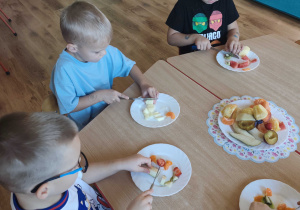 Dzieci siedzą przy stolikach i kroją owoce na sałatkę owocową