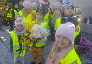 Kazik trzyma kwiaty, które zostaną złożone pod pomnikiem poległych harcerek i harcerzy. Pozostałe dzieci stoją obok.