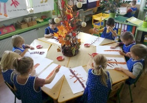 Dzieci siedzą przy okrągłym stole i malują farbami swoje drzewo.
