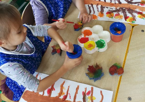Dziewczynki farbami plakatowymi w jesiennych kolorach, maluja gąbkami w kształcie lisci. Robią nimi stemple na swoich pracach przedstawiajacych drzewa.