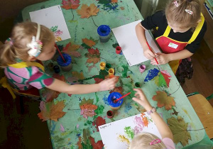 Trzy dziewczynki malują liście, które następnie odciskają na białych kartkach.