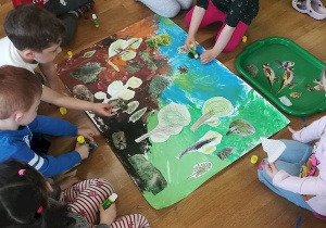 Dzieci tworzą plakat przyklejając elementy przedstawiające rośliny i zwierzęta na tło z kolorami ziemi.