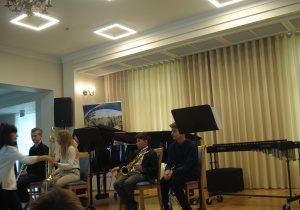 uczniowie Szkoły Muzycznej siedzą na krzesłach na scenie, trzymają w rękach instrumenty: trąbkę, puzon, saksofon i klarnet