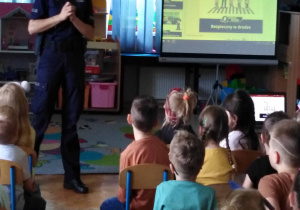 Policjantk stoi w lekkim rozkroku. Dzieci siedzą na krzesełkach.