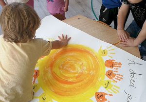 Przedszkolaki tworzą plakat pt. "A dzieci jak promyczki" z odciśniętych dłoni pomalowanych słonecznymi kolorami.