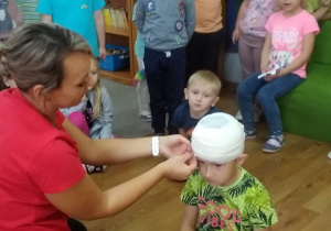 Pani ratownik pokazuje dzieciom, jak należy zabandażować głowę. Maciek siedzi na podłodze z bandażem na głowie.