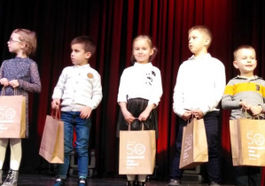 piątka dzieci stoi na scenie i trzyma w rękach nagrody w torbach