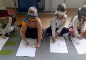 Dzieci siedzą na dywanie z zawiązanymi oczami i rysują na białych kartkach misie.