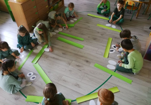 Dzieci siedząc na podłodze robią opaski z zielonych pasków kartonu i kreatywnych patyczków.