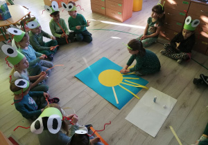 Dzieci przyklejają różnej długości żółte paski (promyczki) na niebieski karton z dużym żółtym kołem(słońcem).