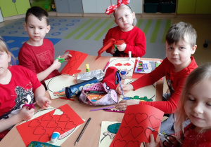 Dzieci ubrane na czerwono siedza przy stolikach i wykonują pracę plastyczną.