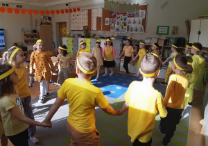 Dzieci ustawione w kole, trzymają się za ręce, tańczą i śpiewają piosenkę.