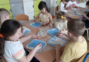 Dzieci siedzące przy stolikach malują papierowe talerzyki farbą plakatową.