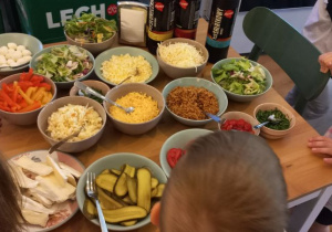Dzieci siedzą przy stole pełnym składników do przygotowania pysznego hot doga: różne rodzaje sera, prażona cebulka, ogórki, sałata, pomidory, kukurydza, szczypiorek
