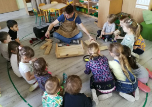 Tata Alicji pokazuje dzieciom siedzącym na elipsie gotowe drewniane elementy, z których zrobią karmnik dla ptaków.