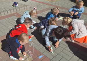 Dzieci rysują kropki kolorową kredą na chodniku przed budynkiem przedszkola.