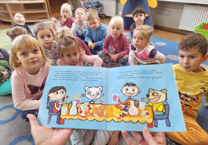 Dzieci słuchają książeczki "Kicia Kocia. Witaminowe przyjęcie", czytanej przez nauczycielkę.