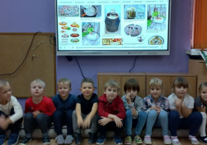 Dzieci siedzą obok siebie w rzędzie, poznają różne rodzaje beczek do kiszenia kapusty