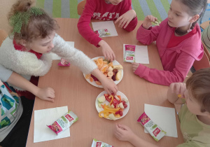 Dzieci siedzą przy stolikach i jedzą warzywa i owoce