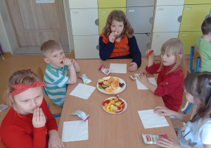 Dzieci siedzą przy stolikach i jedzą warzywa i owoce