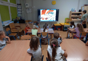 Dzieci siedza na krzesełkach i oglądają na monitorze bajkę edukacyjną na temat dnia kropki